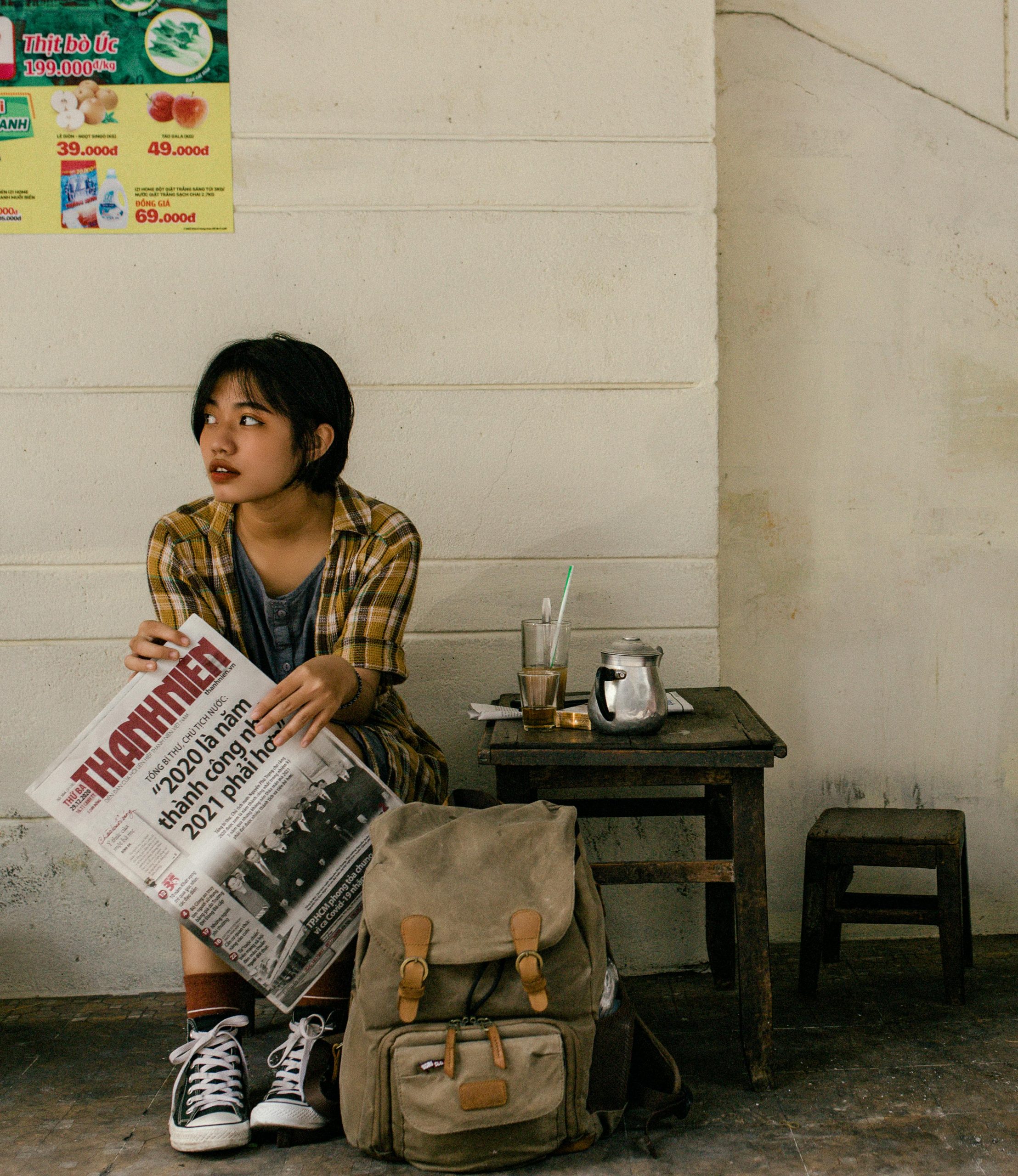 Young woman sitting outdoors with a newspaper and backpack, looking thoughtful.